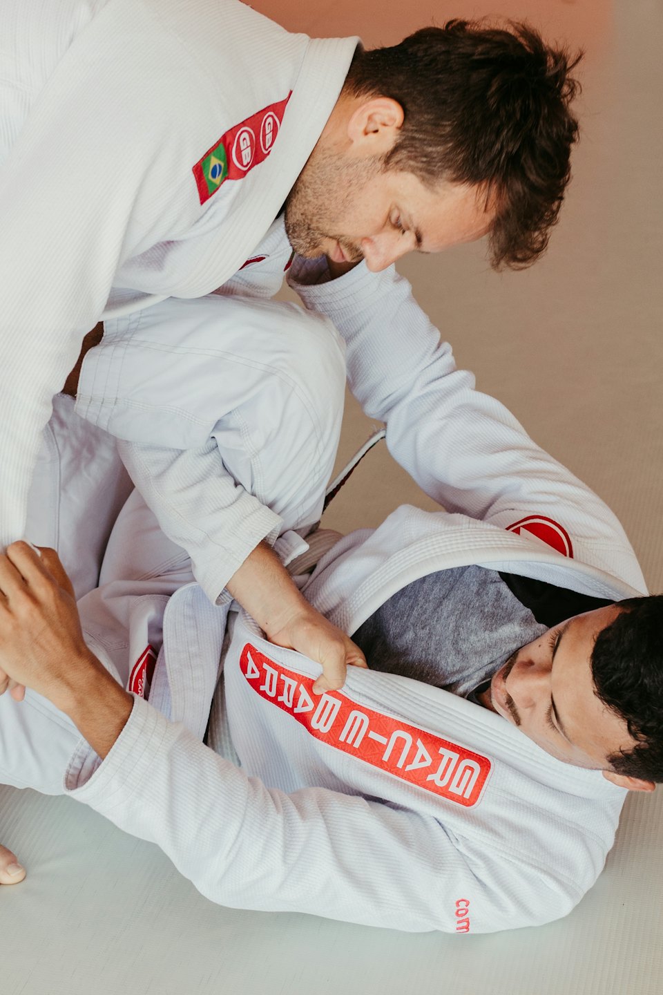 Two men in white gis training Brazilian Jiu-Jitsu on a mat.